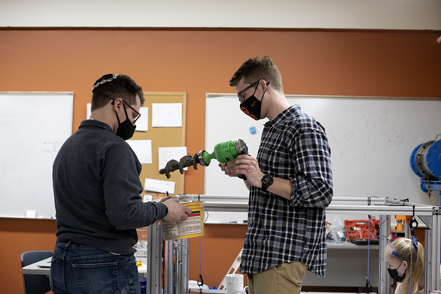 One student holding a drill and one student holding a bucket, working in a engineering lab.