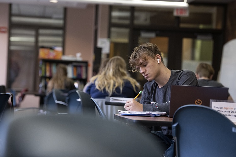 Student with short brown hair, wearing a long sleeve grey shirt and white earbuds is pictured writing in a notebook.