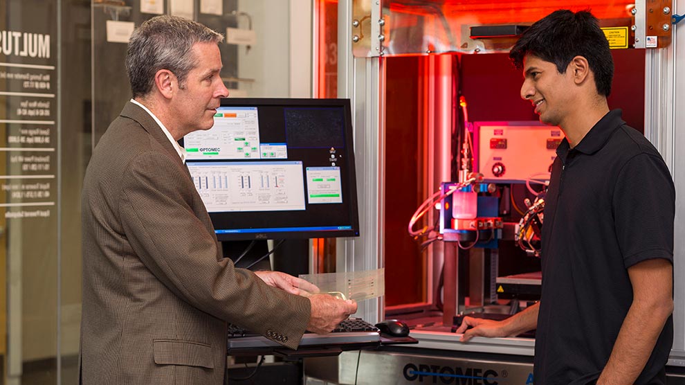 A professor talking to a student in front of an Optomec machine.