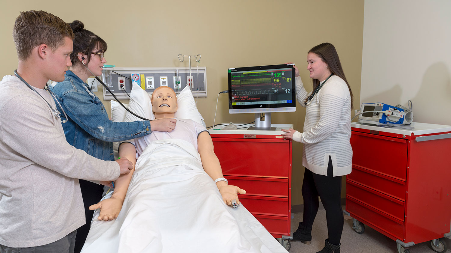 3 students using a stethoscope on a dummy patient and a monitor with data readouts