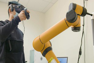 A student wearing a black sweatshirt and virtual reality goggles interacts with a robotic device.