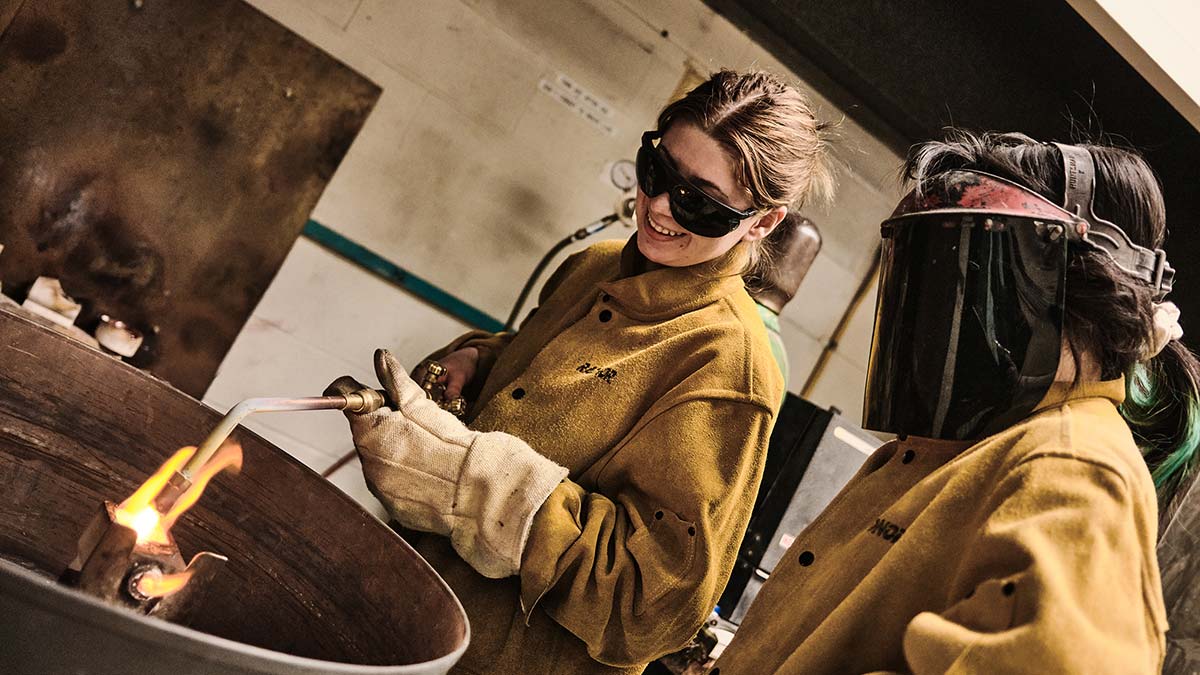 Two students wearing protective leather coats and gloves operate a torch to melt objects for a casting assignment.