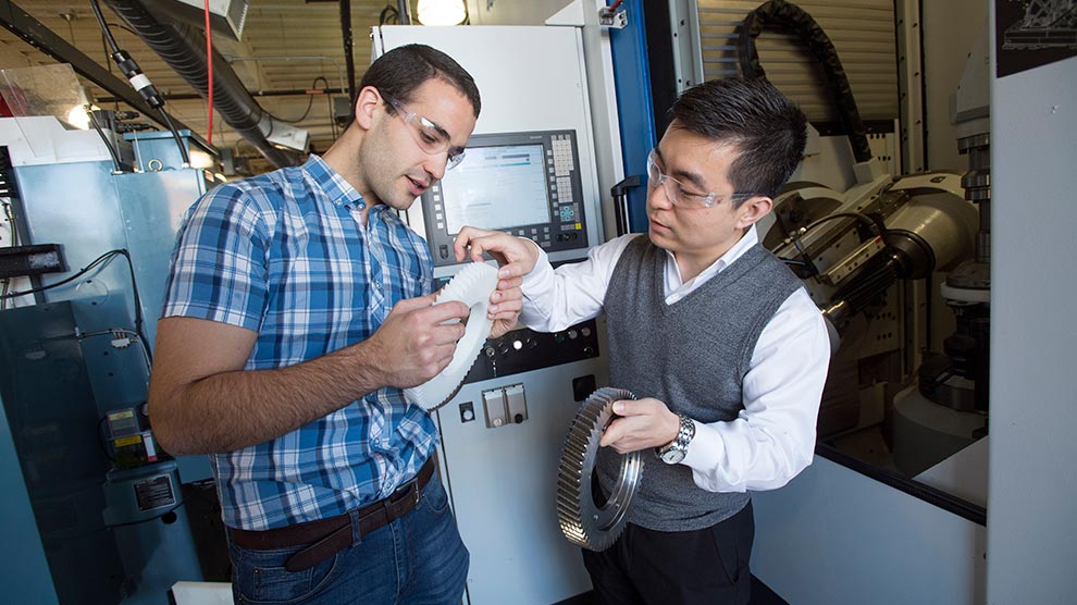 A student in a plaid shirt holding a plastic gear and a faculty member holding a metal gear.