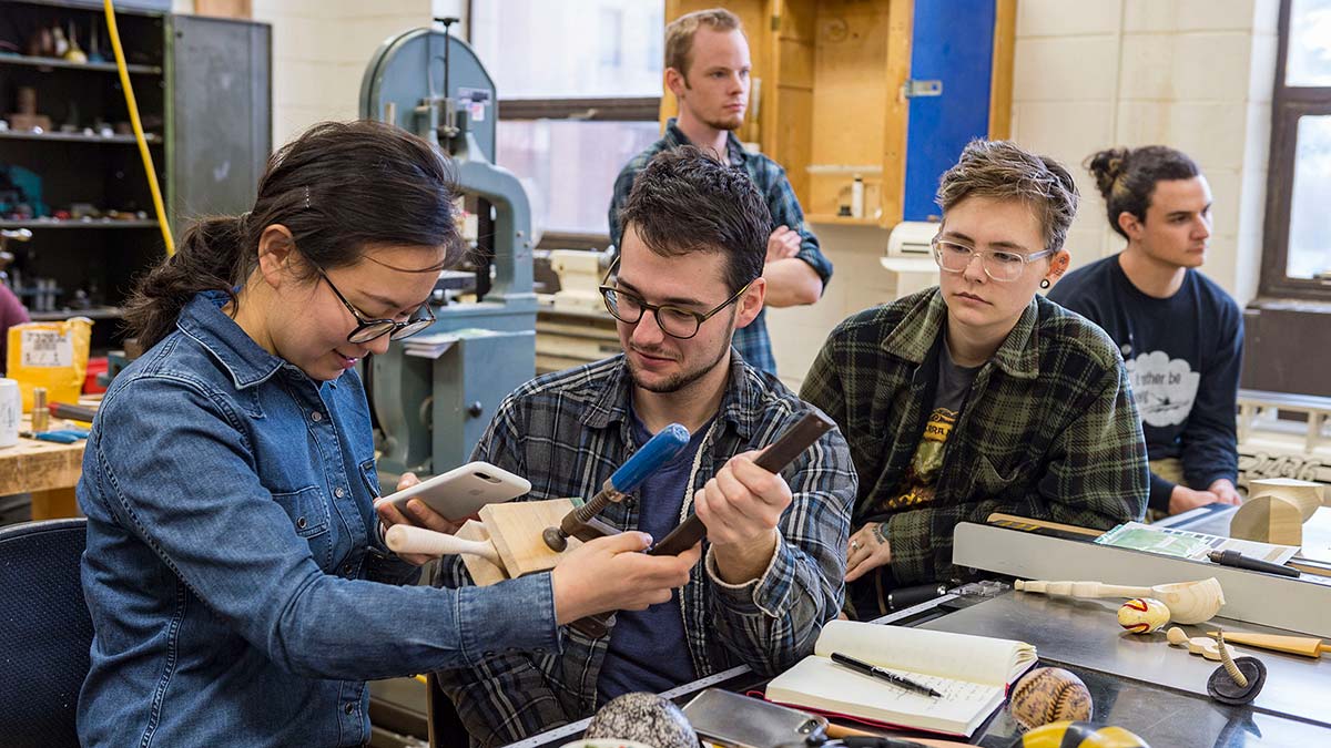 student holds a wooden art piece in a clamp while another person takes a photo of it.