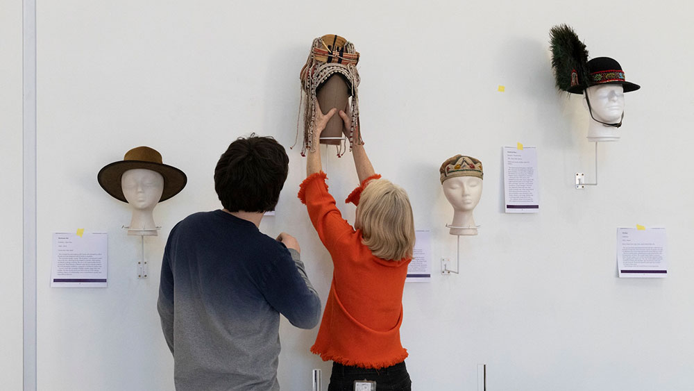 Two people placing headwear on display in a gallery