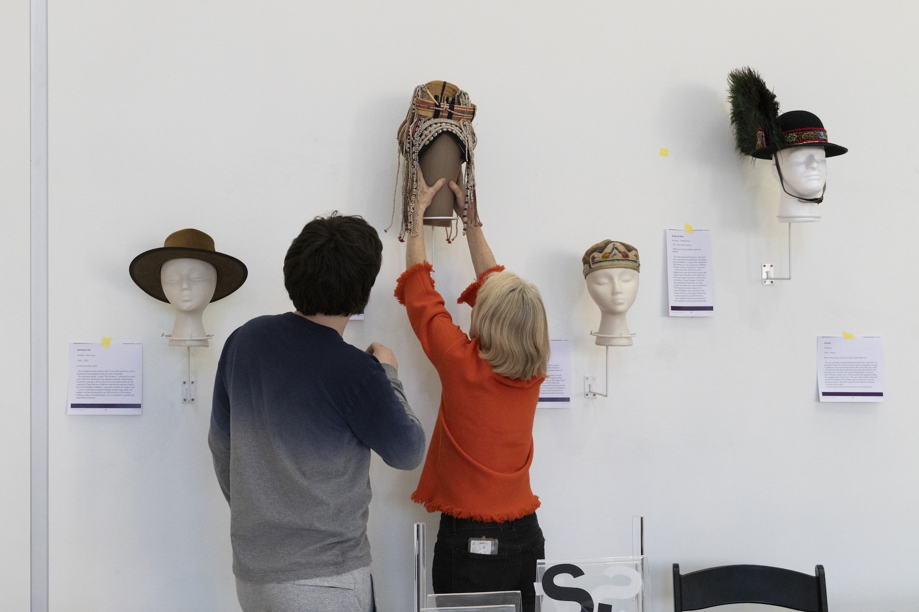 Two people placing headwear on display in a gallery