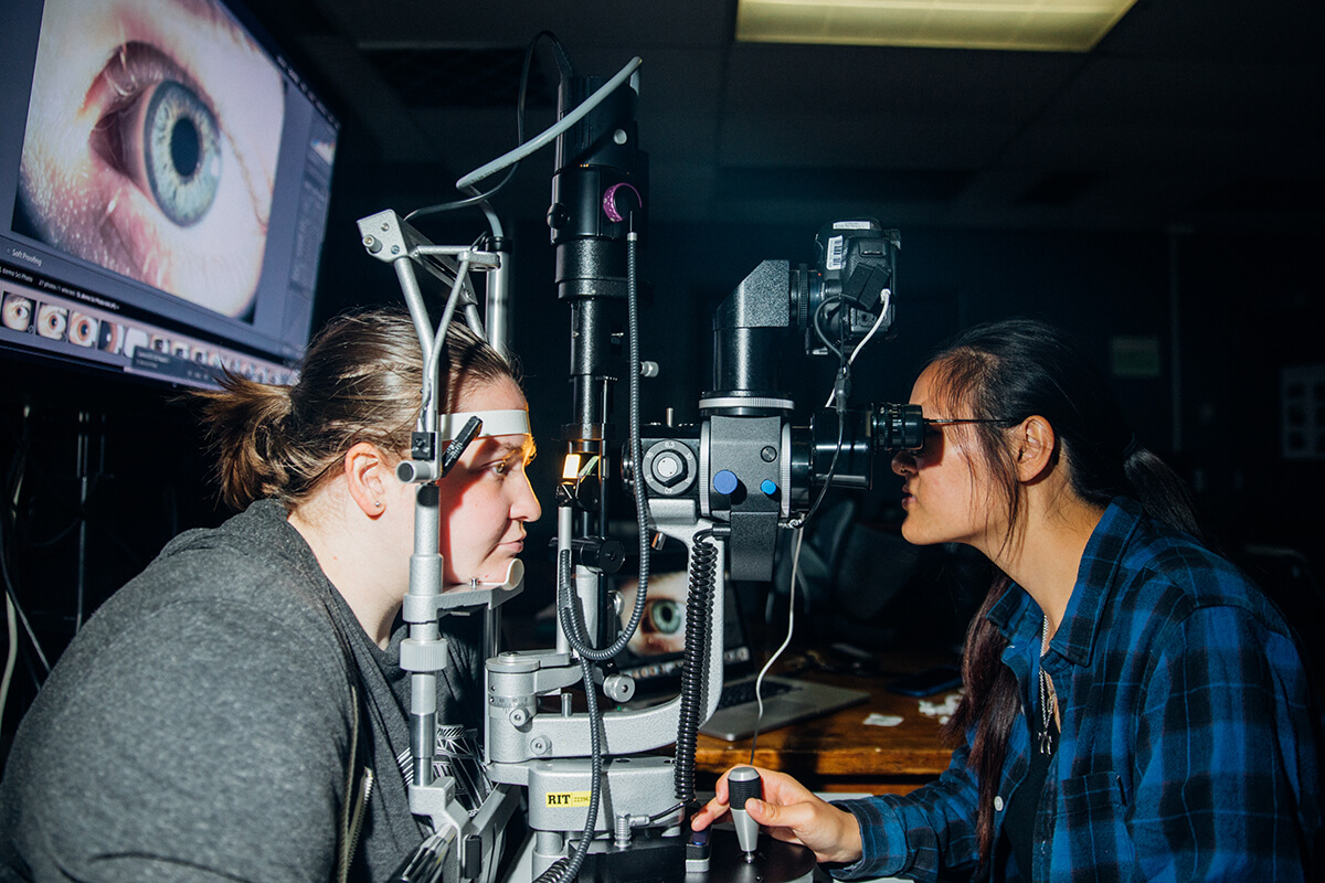a student looks through a machine commonly found in eye doctor offices, as another student acts as the patient.