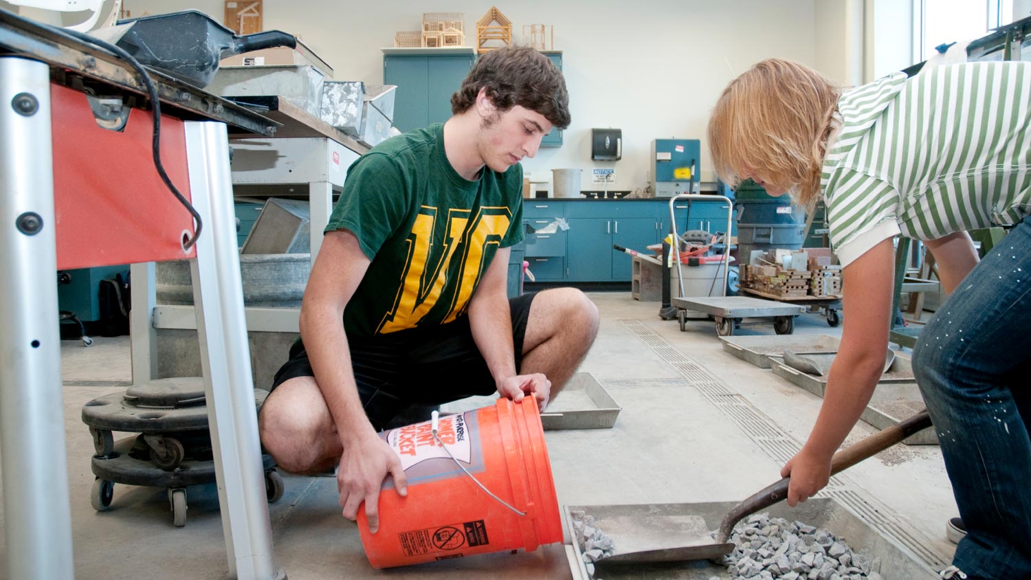 Two students scooping small stones into a 5 gallon bucket