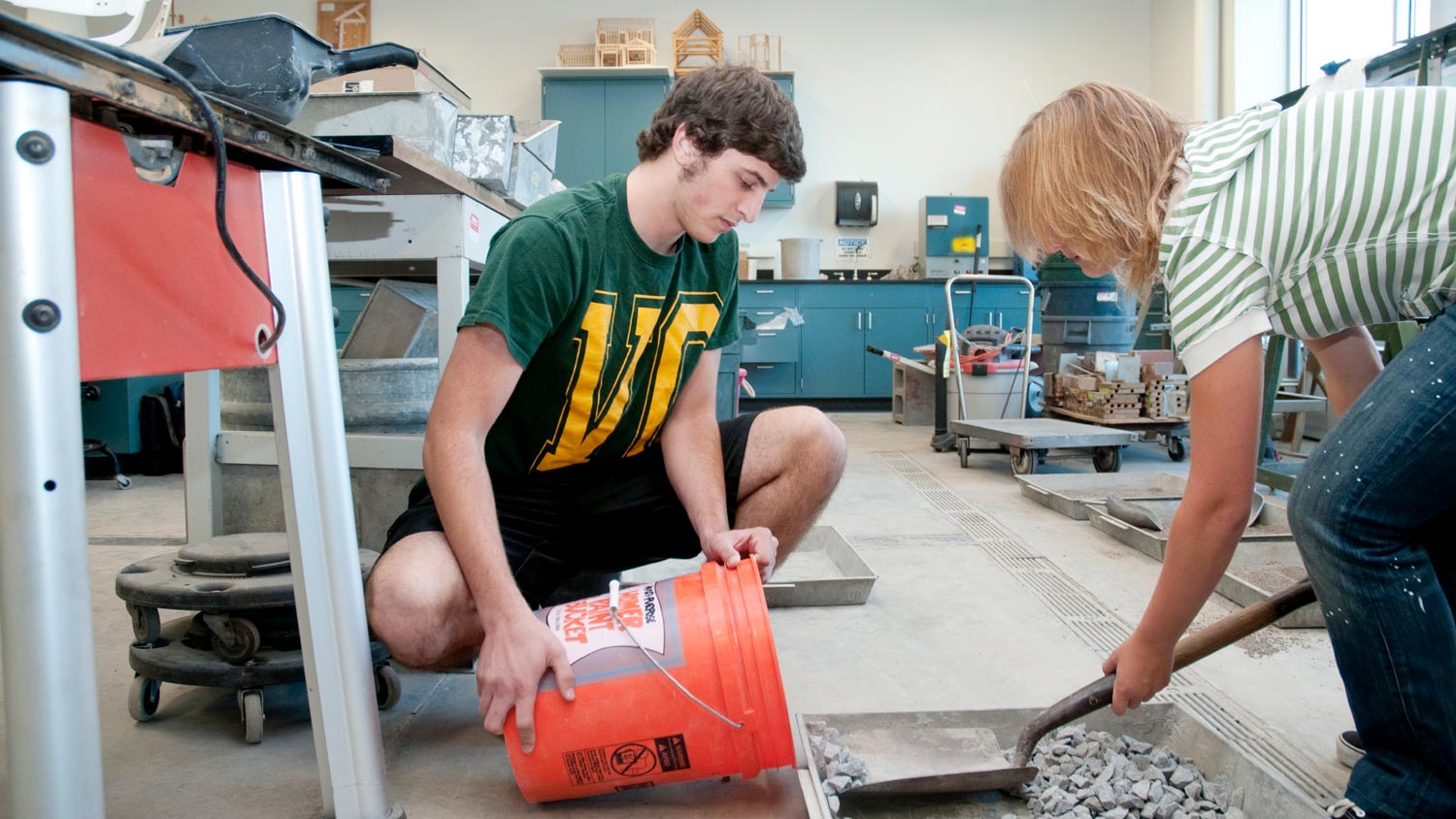 Two students scooping small stones into a 5 gallon bucket.