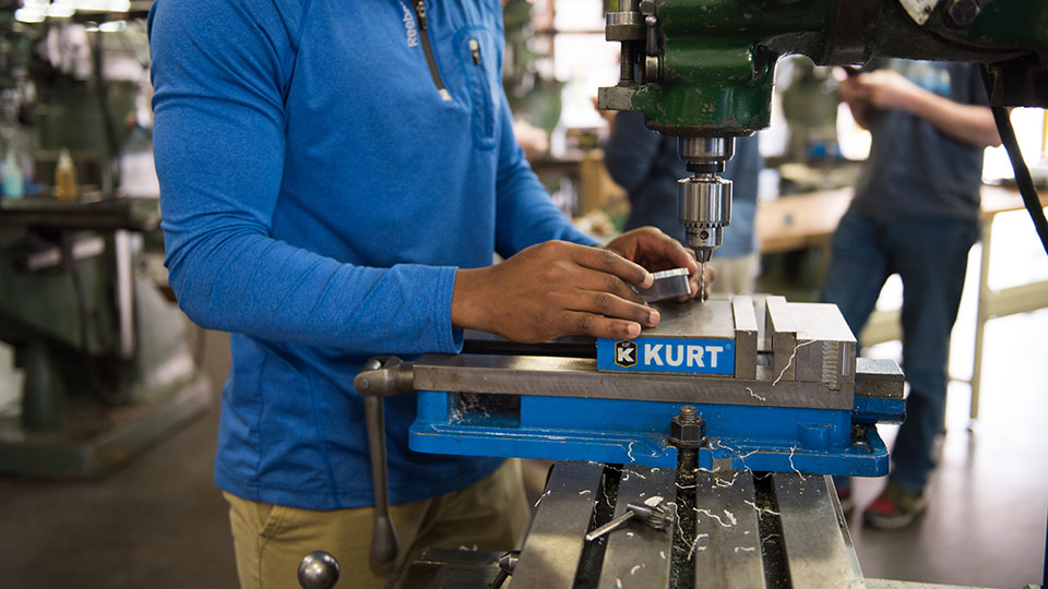 Student wearing a blue shirt working with a drill press.