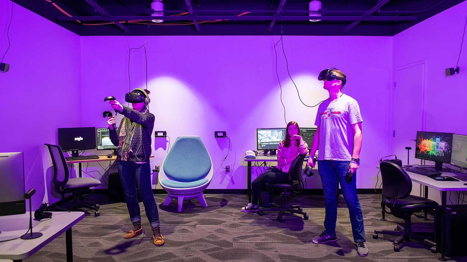 Two students stand wearing VR goggles and holding controllers in room with chairs and computers