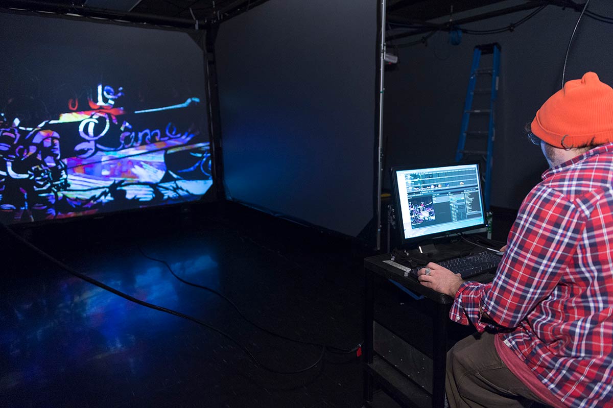 student works on a laptop as a scene is projected onto a large screen in a dark room.