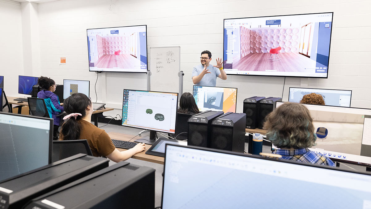 professor gesturing at front of computer lab, with two large T Vs displaying a chair in a room.
