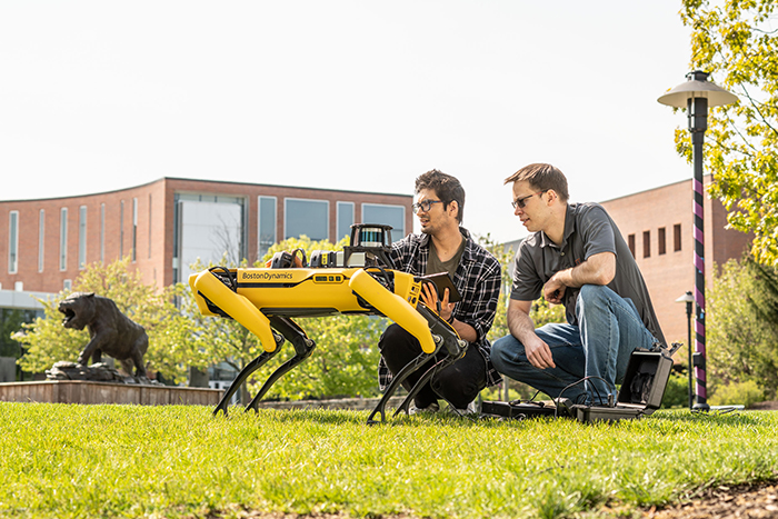 Two people squatted down, looking at Boston Dynamics robot, Spot who is yellow and shaped like a dog.