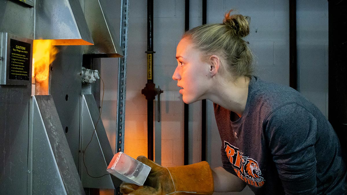 a student looks into a kiln while holding a ceramic brick with a protective leather glove.