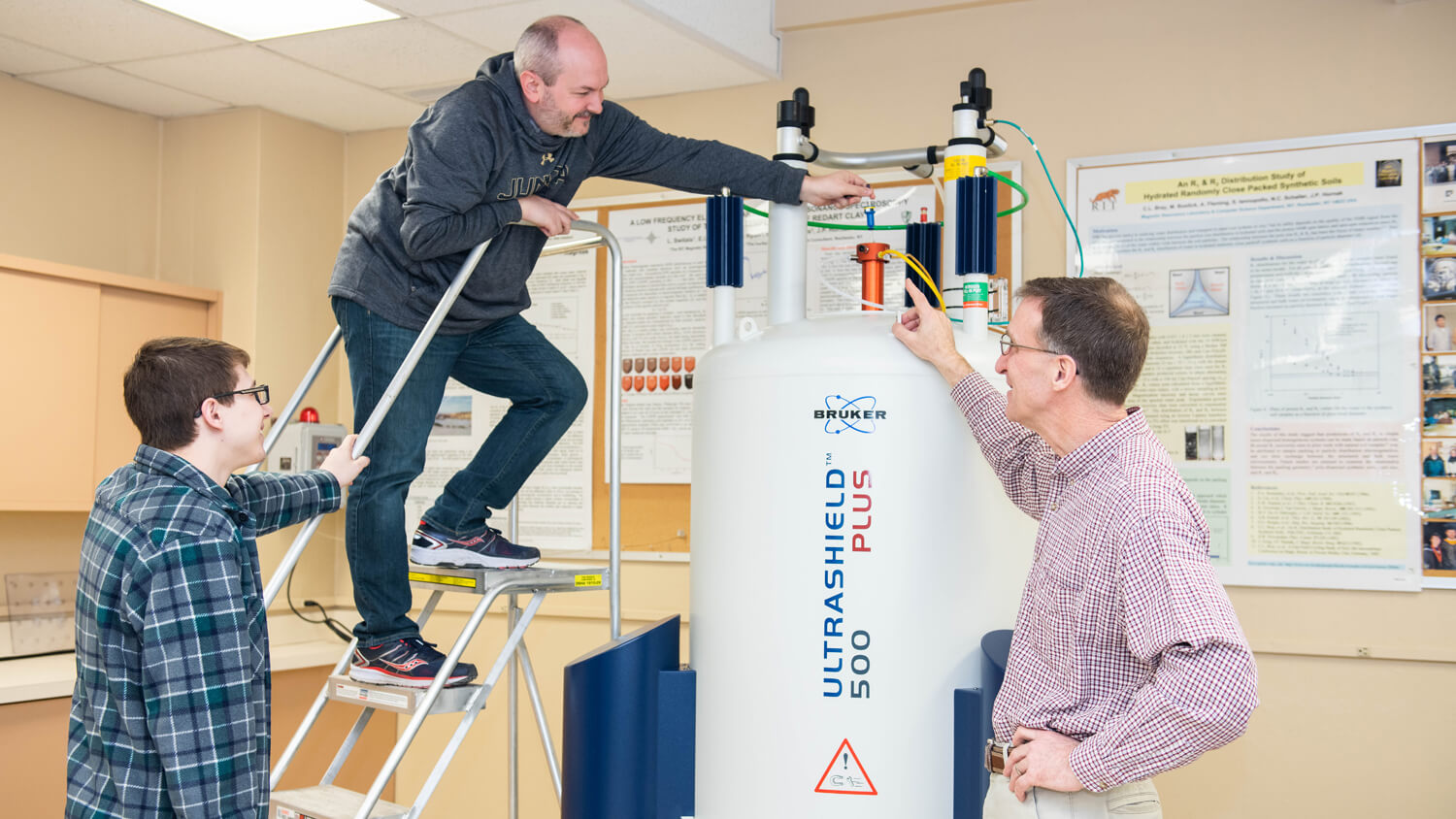 a man climbs a stepstool to reach the top of a Bruker Ultrashield 500 spectrometer, a large tank, as two others look on.