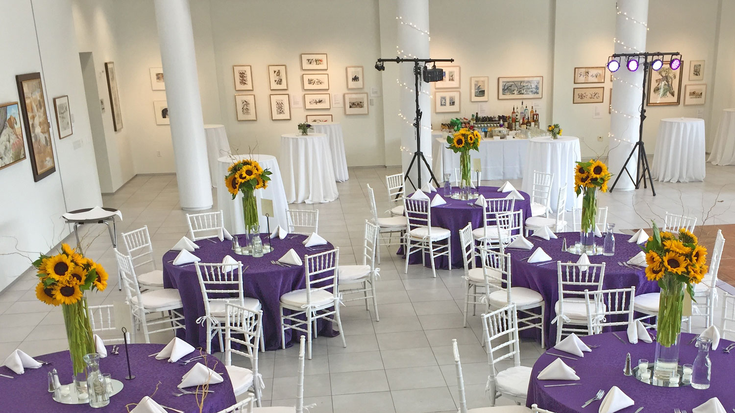 The university gallery space set up with purple table-clothed tables with sunflower bouqets.