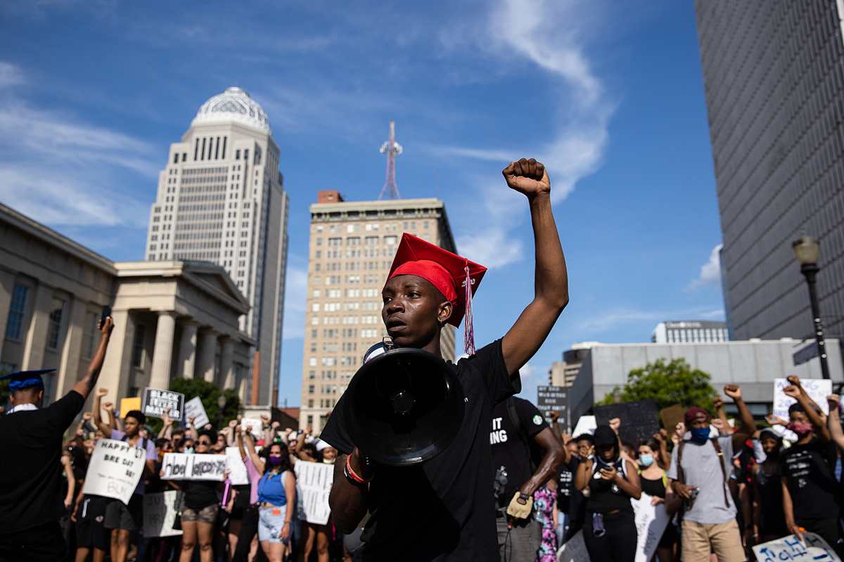 A boy wearing a graduating cap raises his left arm in the air, surrounded by a crowd of fellow protestors.