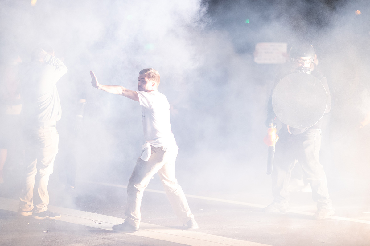 A man, surrounded by a white fog, motions to a crowd of fellow protestors.