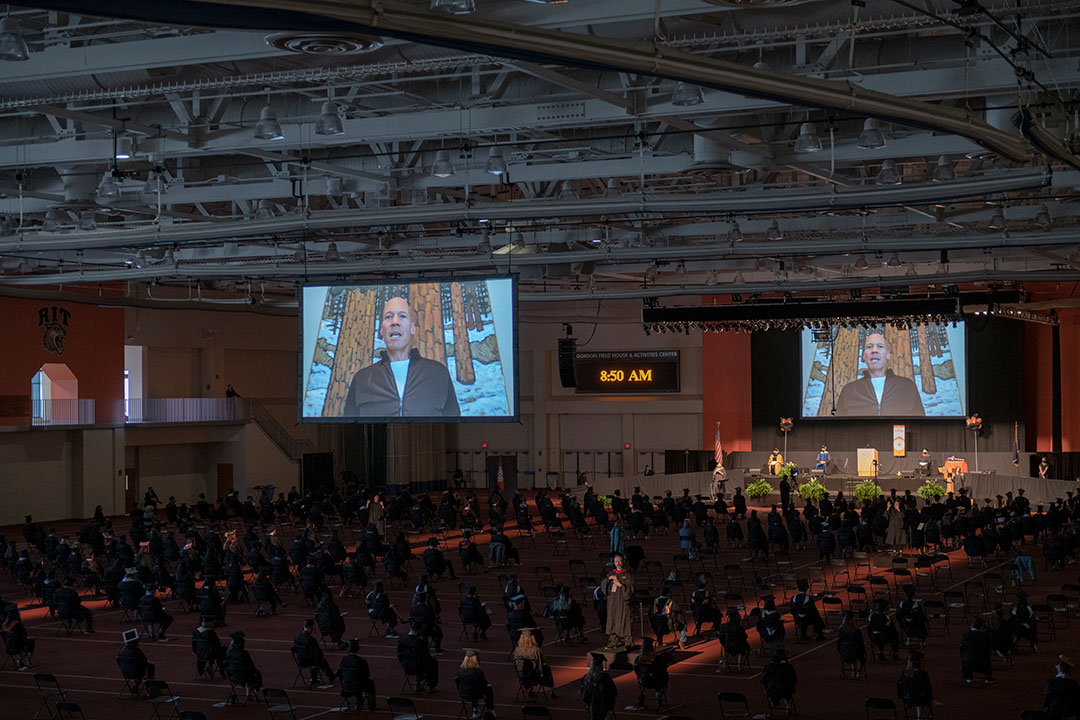 Overhead view of field house with graduates seated six feet apart in chairs on the floor and large screens showing the speaker.