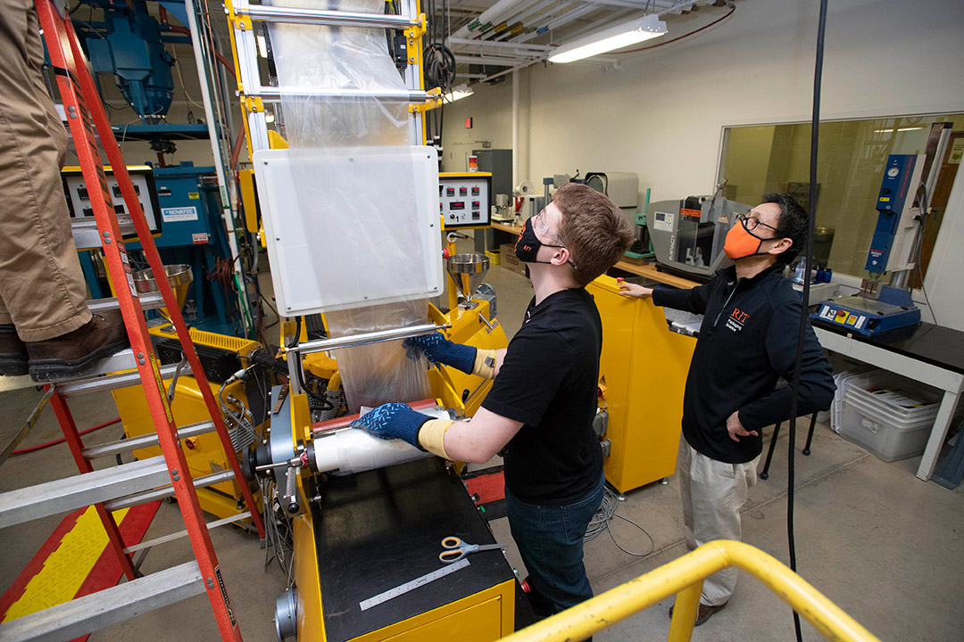 student and professor watch a packaging machine processing plastic wrap.