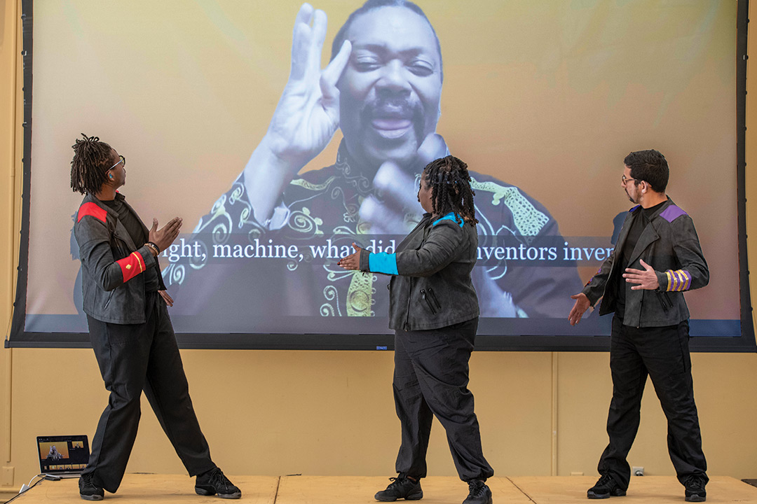 three people turned toward a projector screen behind them showing a man and subtitles.