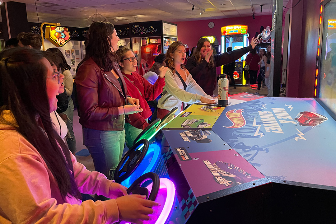 five college students playing a video game with a steering wheel controller.