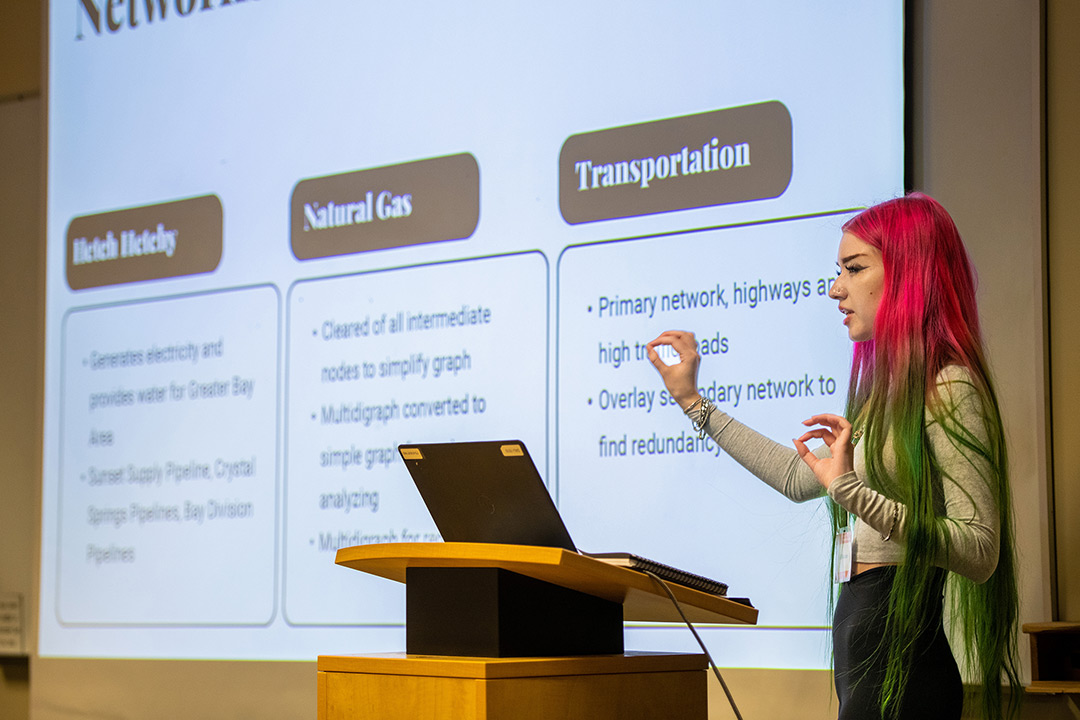 Celia Rickson stands on stage at a podium giving a PowerPoint presentation on her research. She has pink and green hair and is wearing a light green long sleeve shirt with black pants.