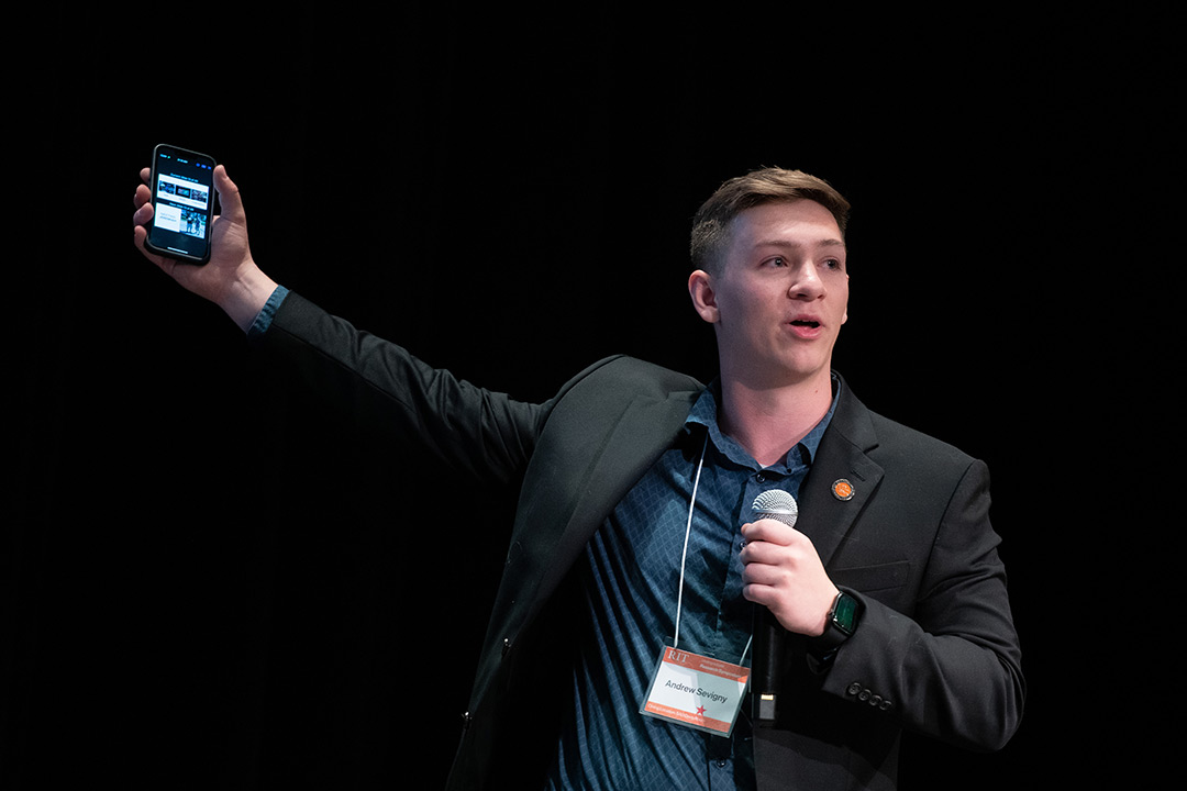 Keynote speaker Andrew Sevigny stands on stage with a microphone in one hand and an iPhone in the other. He is wearing a blue button up shirt and a black blazer with an RIT pin on the lapel.