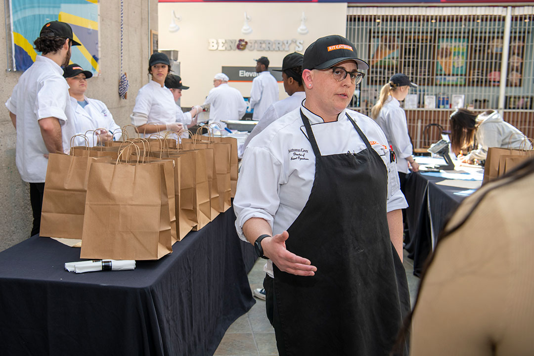 person wearing chef coat and apron talking to another person.