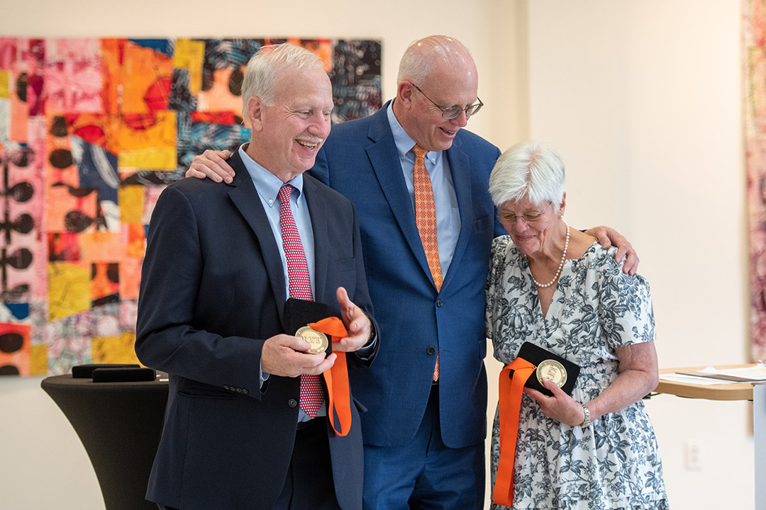 man standing in between another man and a woman, with his arms around their shoulders as they hold R I T medallions.