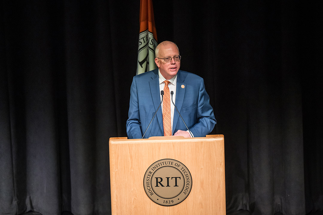 R I T president wearing a blue suit speaking at a podium on a stage next to the R I T flag.