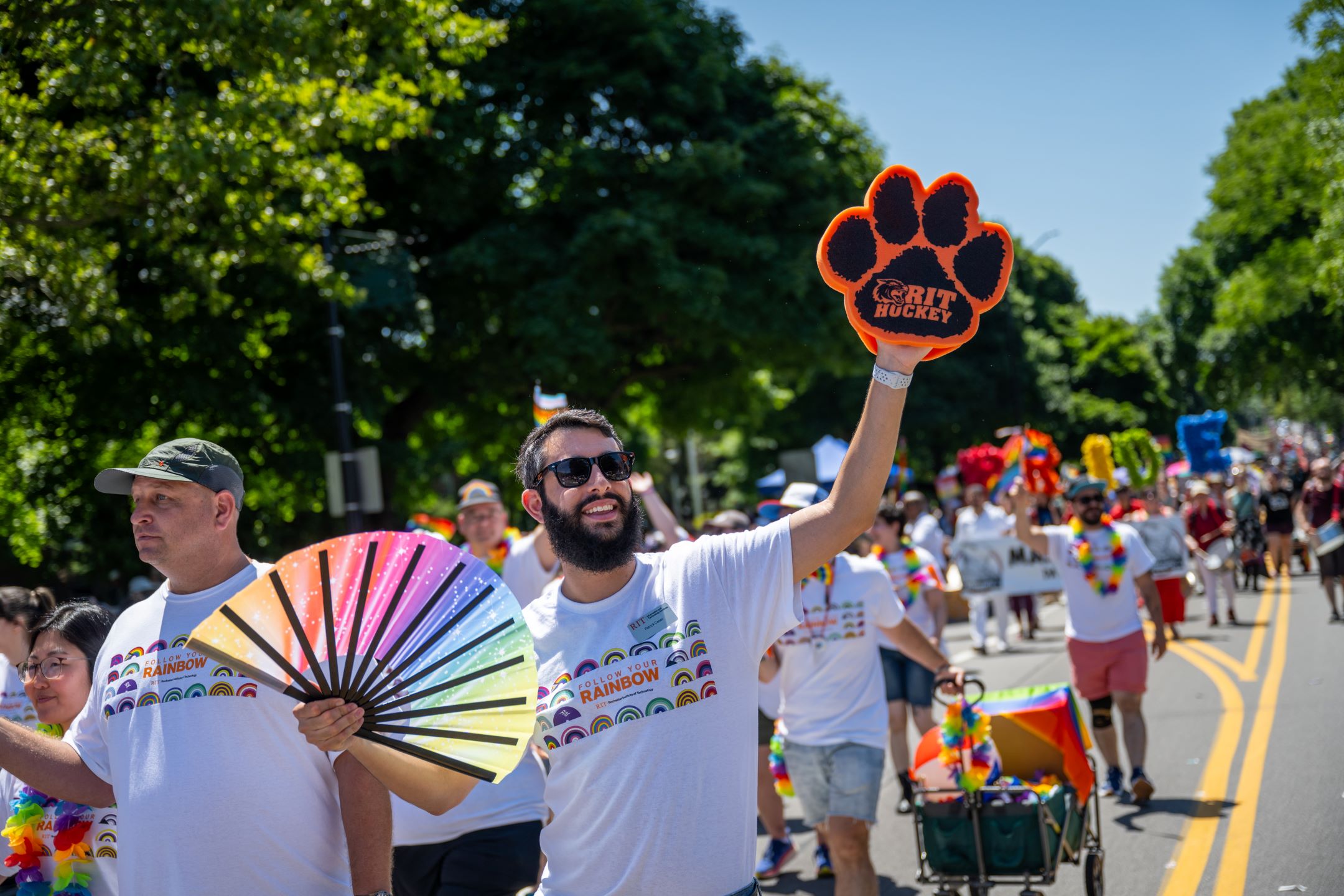 Man with beard walks in parade waving a rainbow fan and with a orange foam tiger paw. 