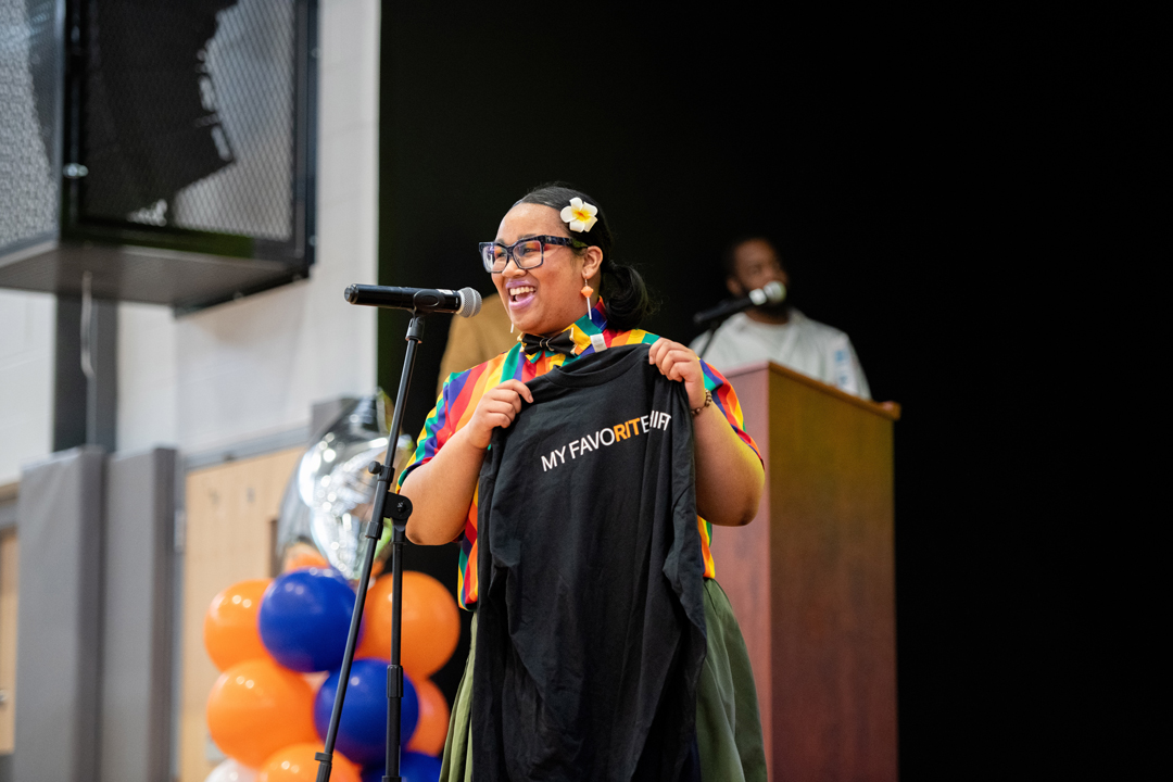 A student stands in front of a microphone on a stage holding a black t-shirt