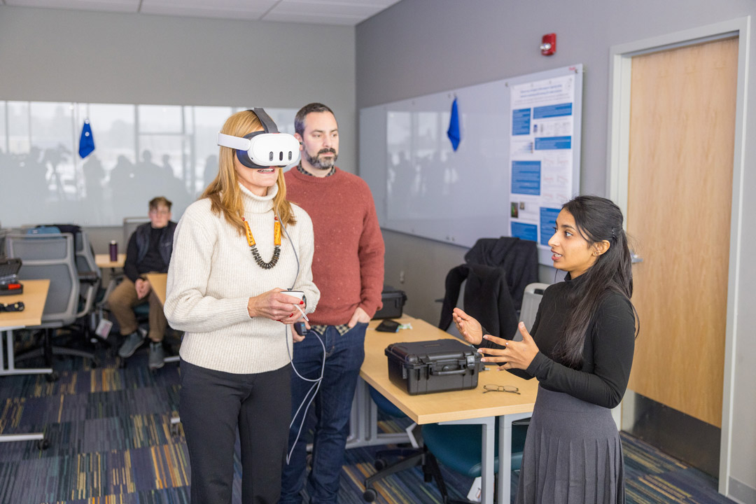 seeral people stand in a classroom environment. One of them has a V R Headset on.