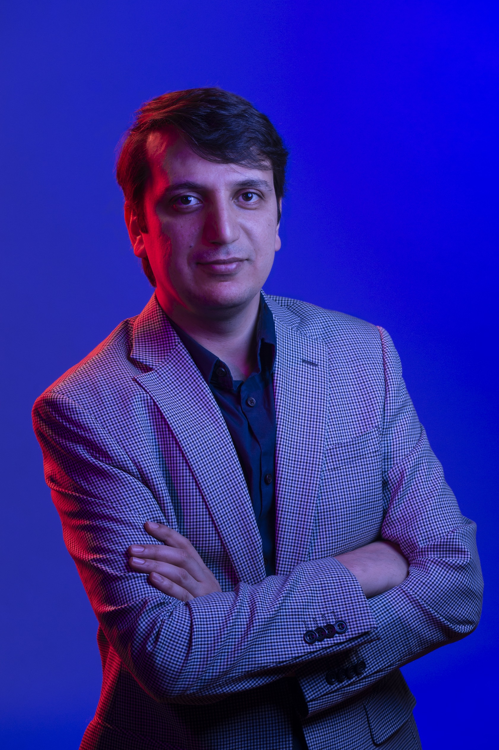 A man in a sportjacket with his arms crossed in front of a blue background