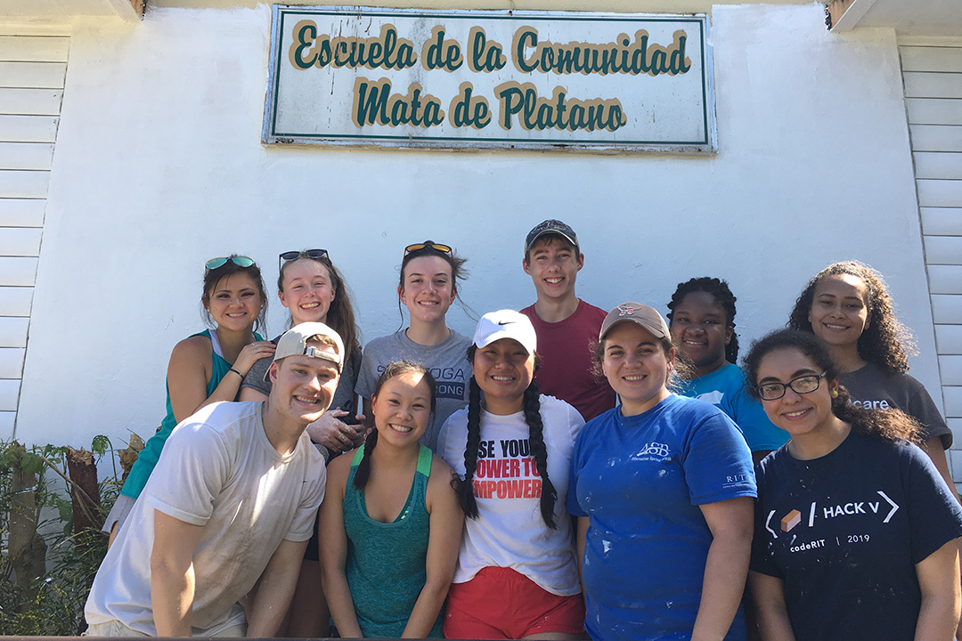 Group of students stands outside building with sign that reads: Escuela de la Comunidad Mata de Platana