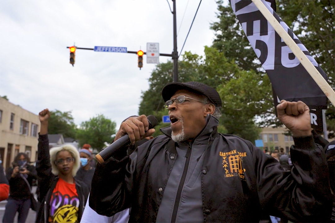 man standing in an intersection speaking into a microphone and holding up his fist.