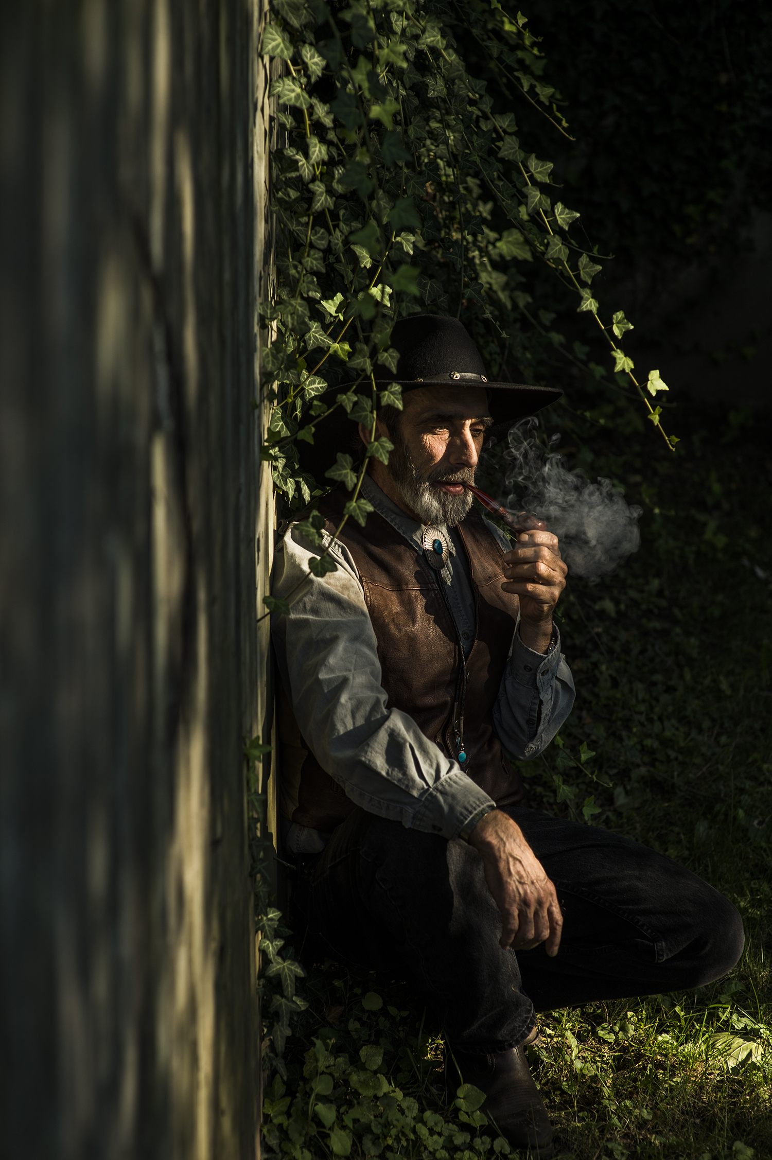 Man in cowboy garb leans against a wall smoking a pipe