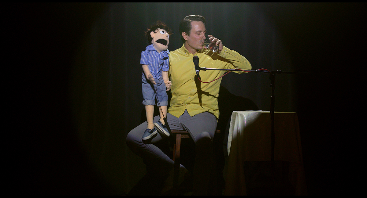 A screenshot from the Teddy and Tater film, with a ventriloquist and his puppet on stage.
