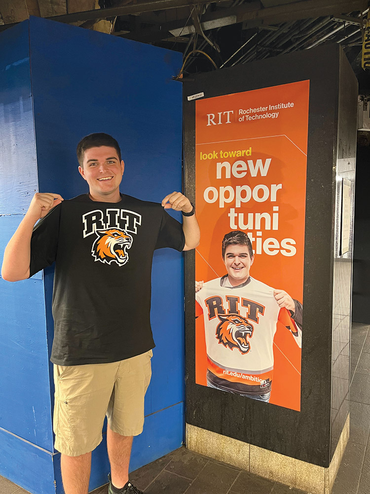 college student standing next to an ad hanging on a subway wall that features his picture.