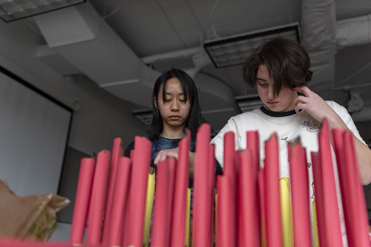 Two students place rolled-up book pages on a grid of pegs. 