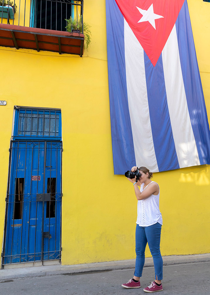 student taking phot in Cuban street with large Cuban flag hanging from building.