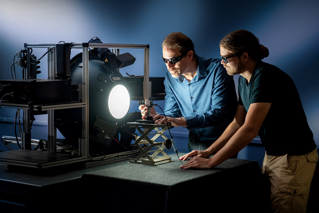 two researchers wearing sunglasses in a dark room, setting up equipment with a bright light.