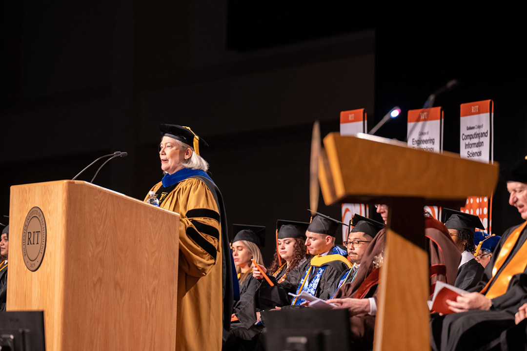 woman in graduation regalia speaking at podium.