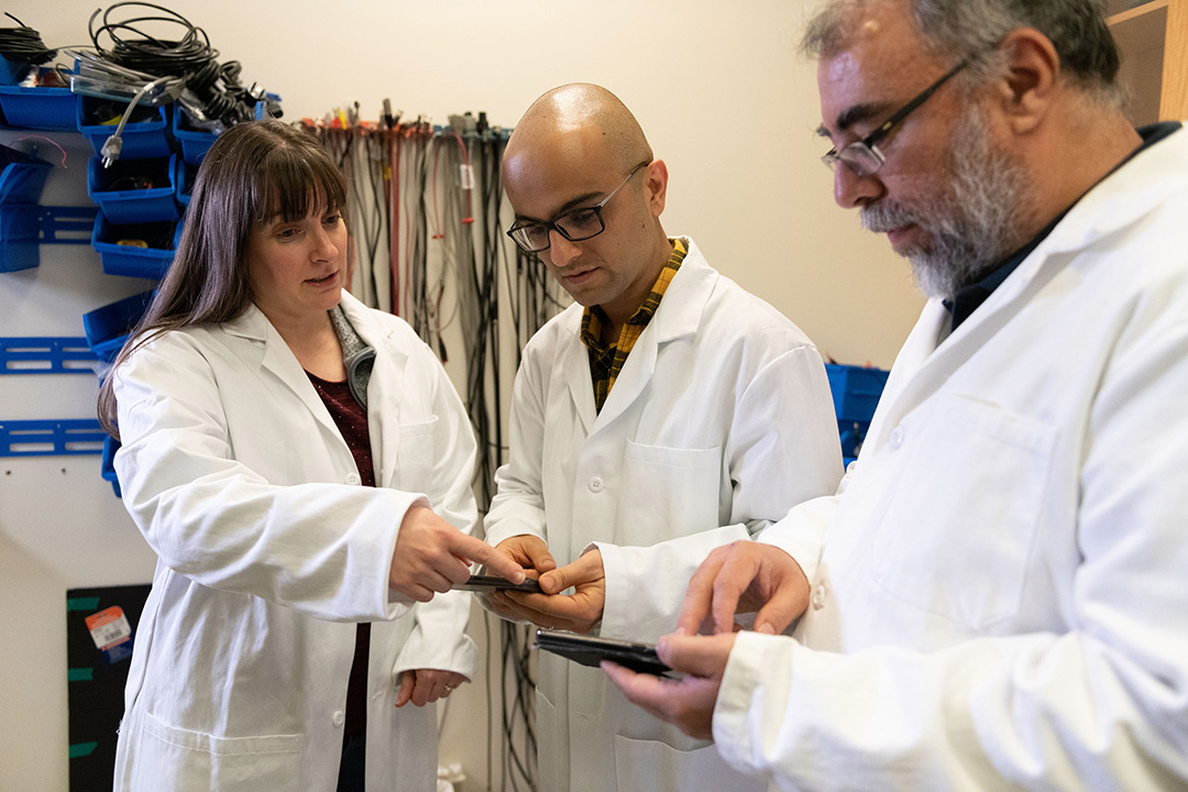 Three researchers wearing lab coats stand pointing at smartphones.