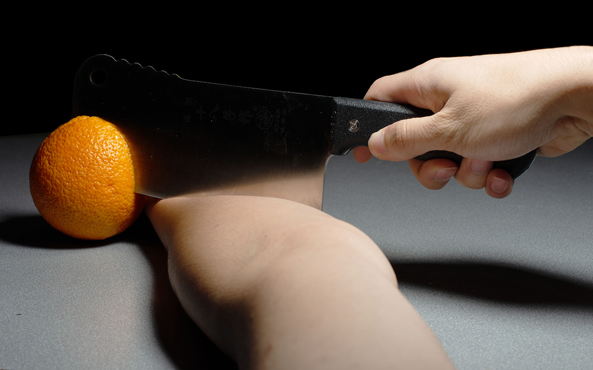 A knife slices through an orange and an arm.