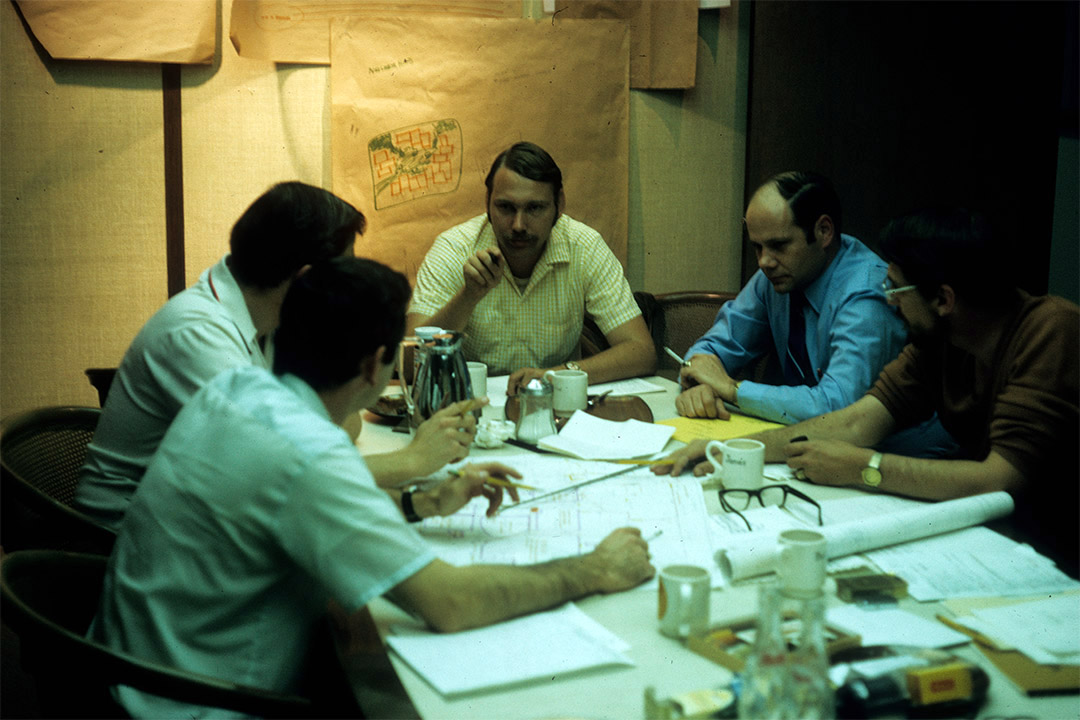 five men sitting at a table looking at maps and blueprints.