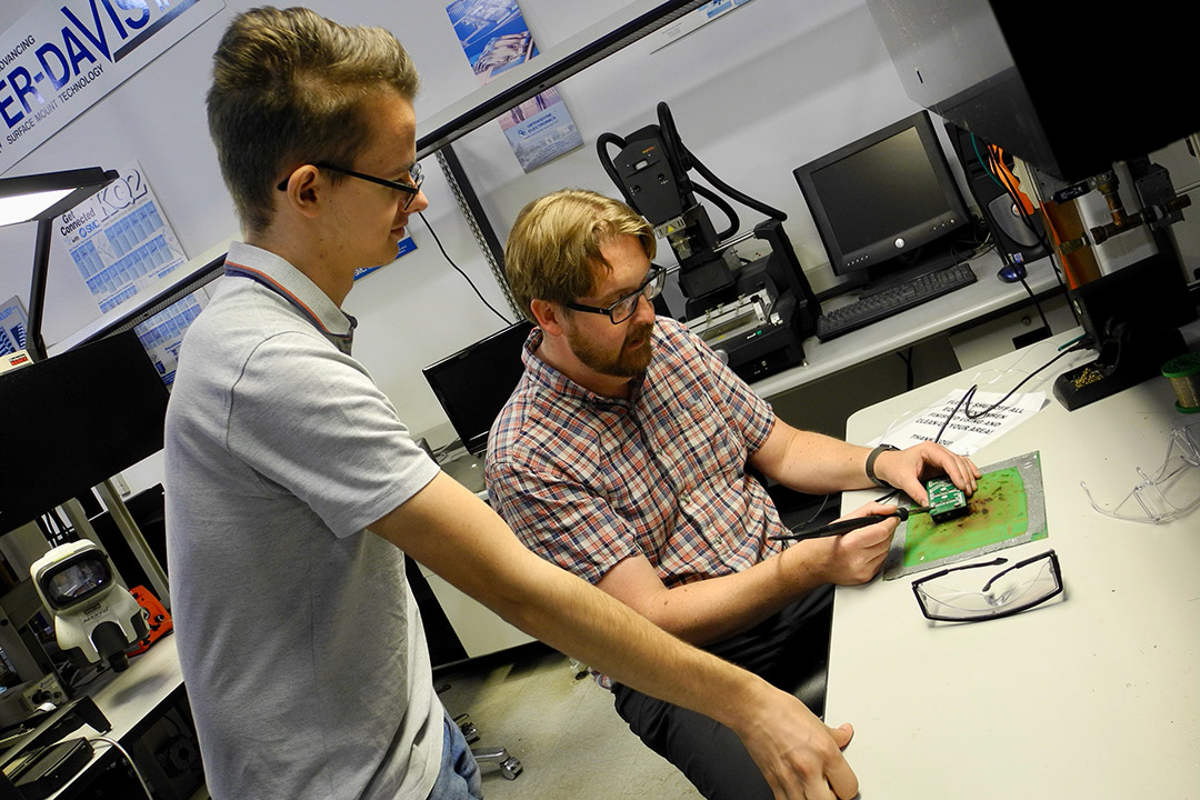 college student watching a professor solder a circuit board.