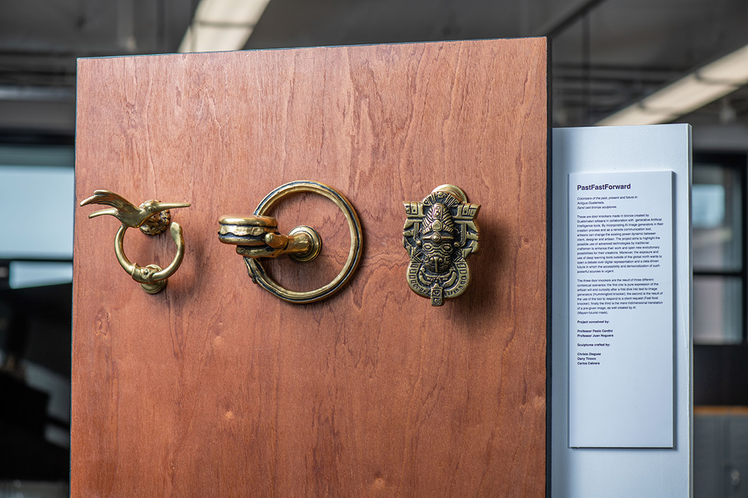 three bronze doorknockers mounted on a door, the first with a hummingbird design, the middle with a cheeseburger design, the third with an ancient Mayan inspired design. 